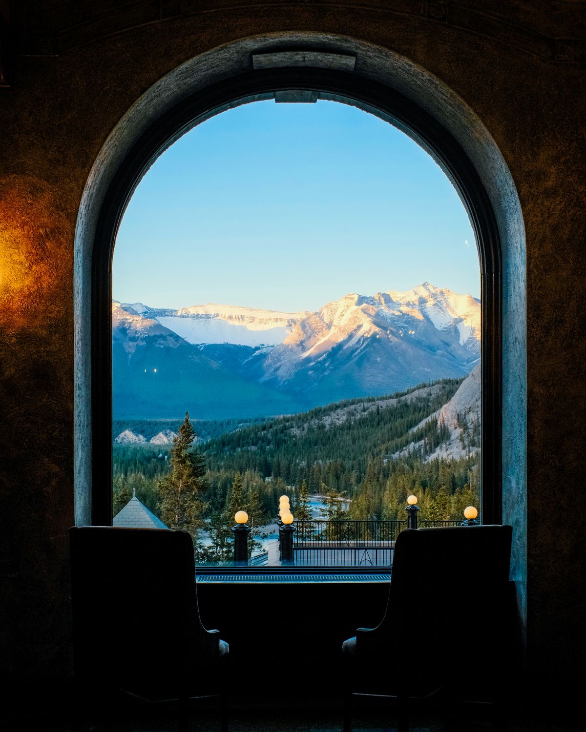 Two armchairs before an arched hotel window looking out on snow-lit mountains at dusk.
