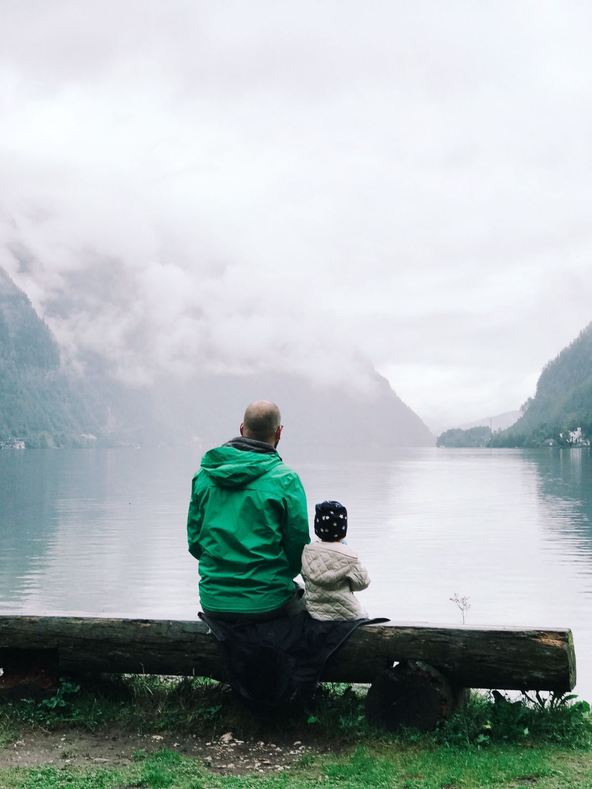 An adult and a child sitting on a log bench at the edge of a misty mountain lake.